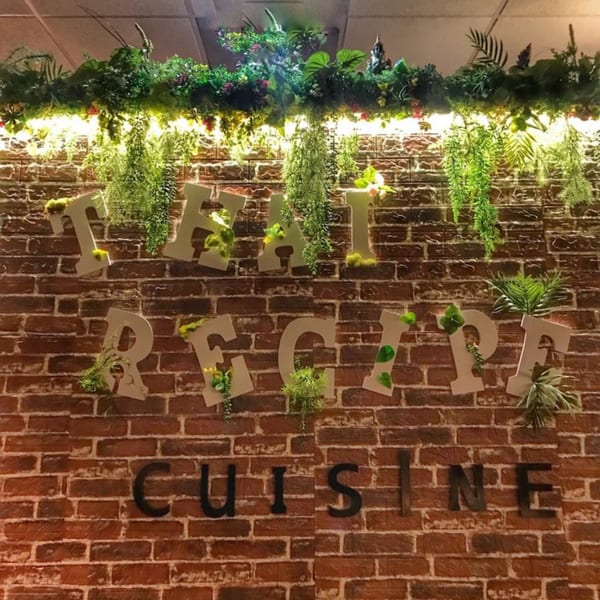 Thai Recipe Cuisine sign on a brick wall, adorned with green plants and warm lighting.