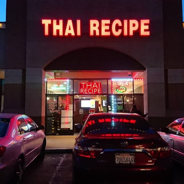 Exterior of Thai Recipe restaurant at night, with bright red neon signs and parked cars reflecting the light.